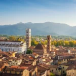 Vista aerea sui tetti in terracotta, i campanili e le chiese romaniche di Lucca con le colline toscane sullo sfondo.