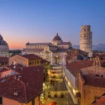 Vista panoramica al tramonto di Piazza dei Miracoli a Pisa con il Battistero, il Duomo e la Torre Pendente illuminata.