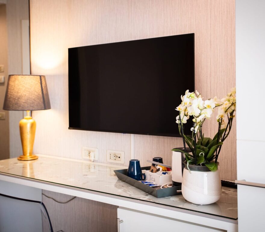 Hotel room desk area with a golden table lamp, wall-mounted flat-screen TV, and a potted white orchid against a neutral wall.